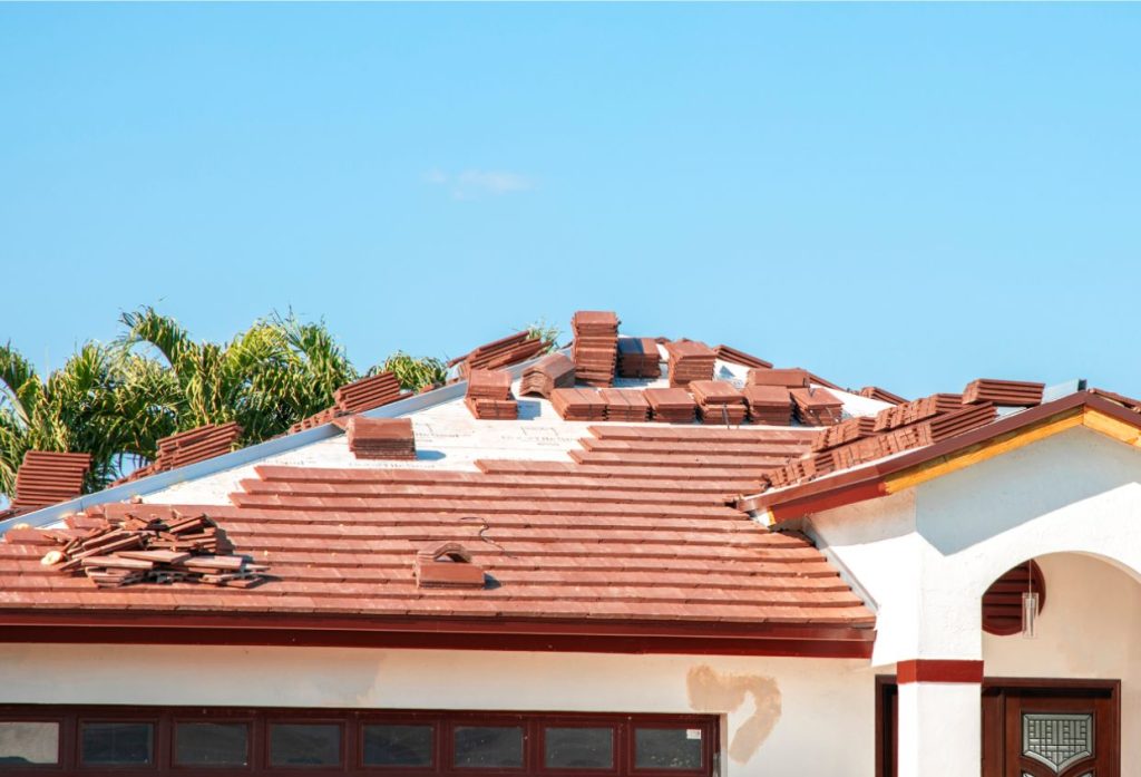 A Spanish-style tile roof is undergoing repair, with sections of red tiles removed and replacement tiles stacked across the roof.