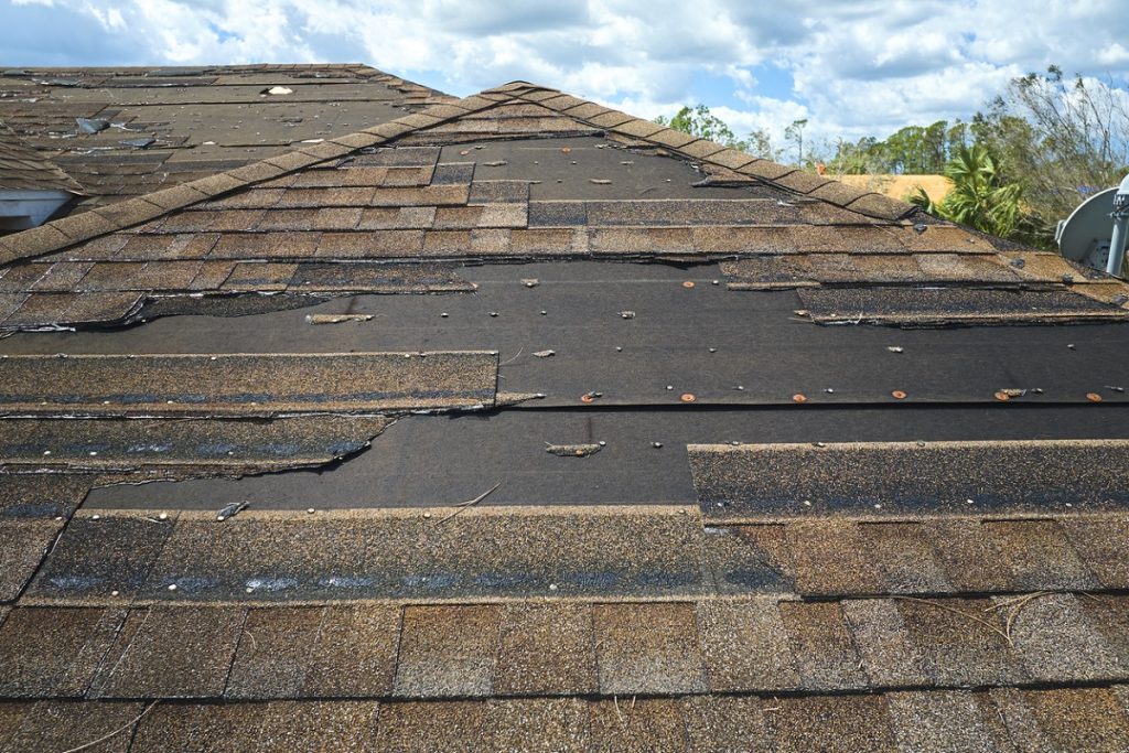 Damaged house roof with missing and torn shingles, exposing the underlying roofing felt.